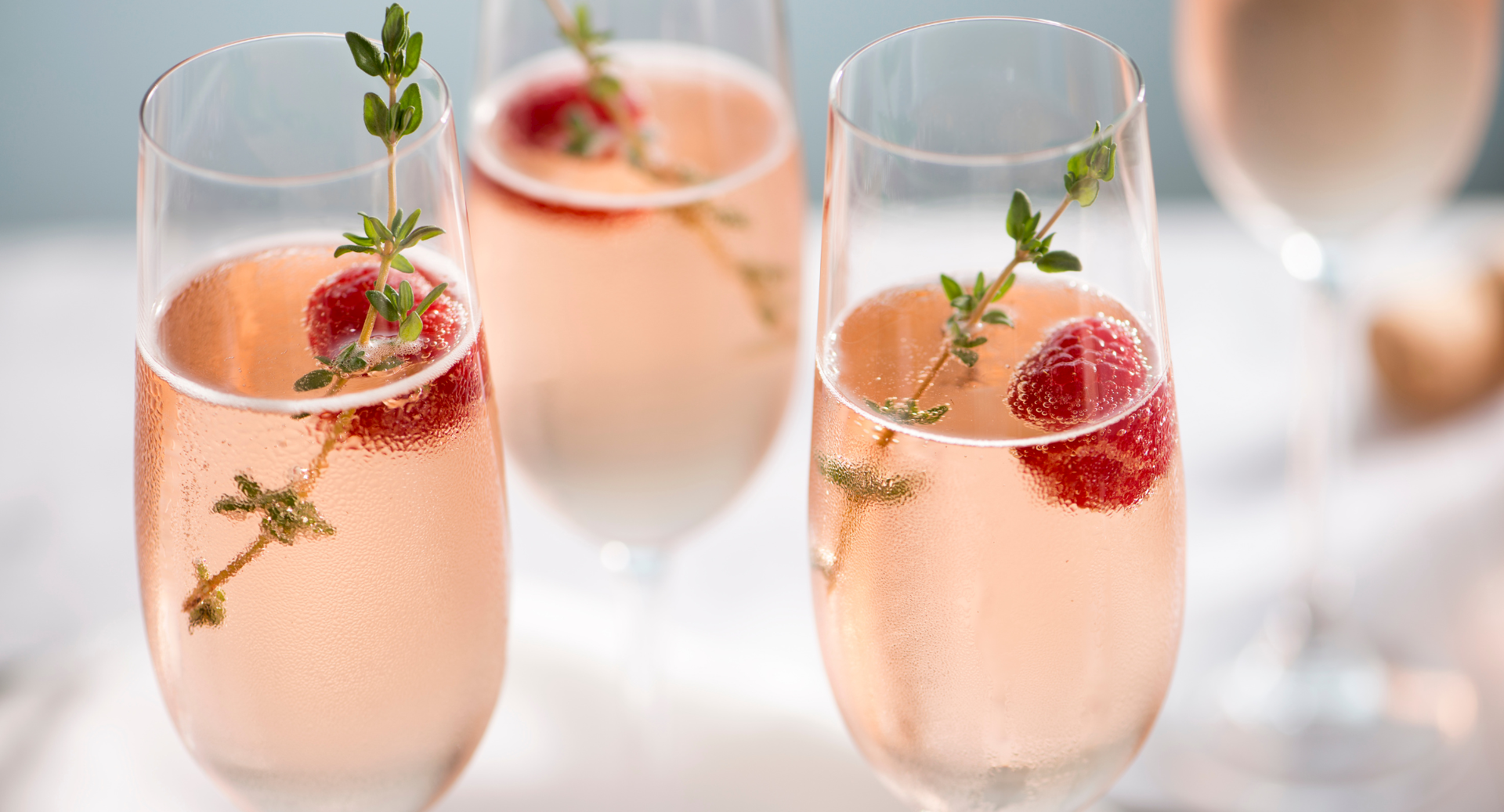 Sparkling peach ros&eacute; glasses with raspberries, thyme garnish, and delicate bubbles on a bright, festive table.
