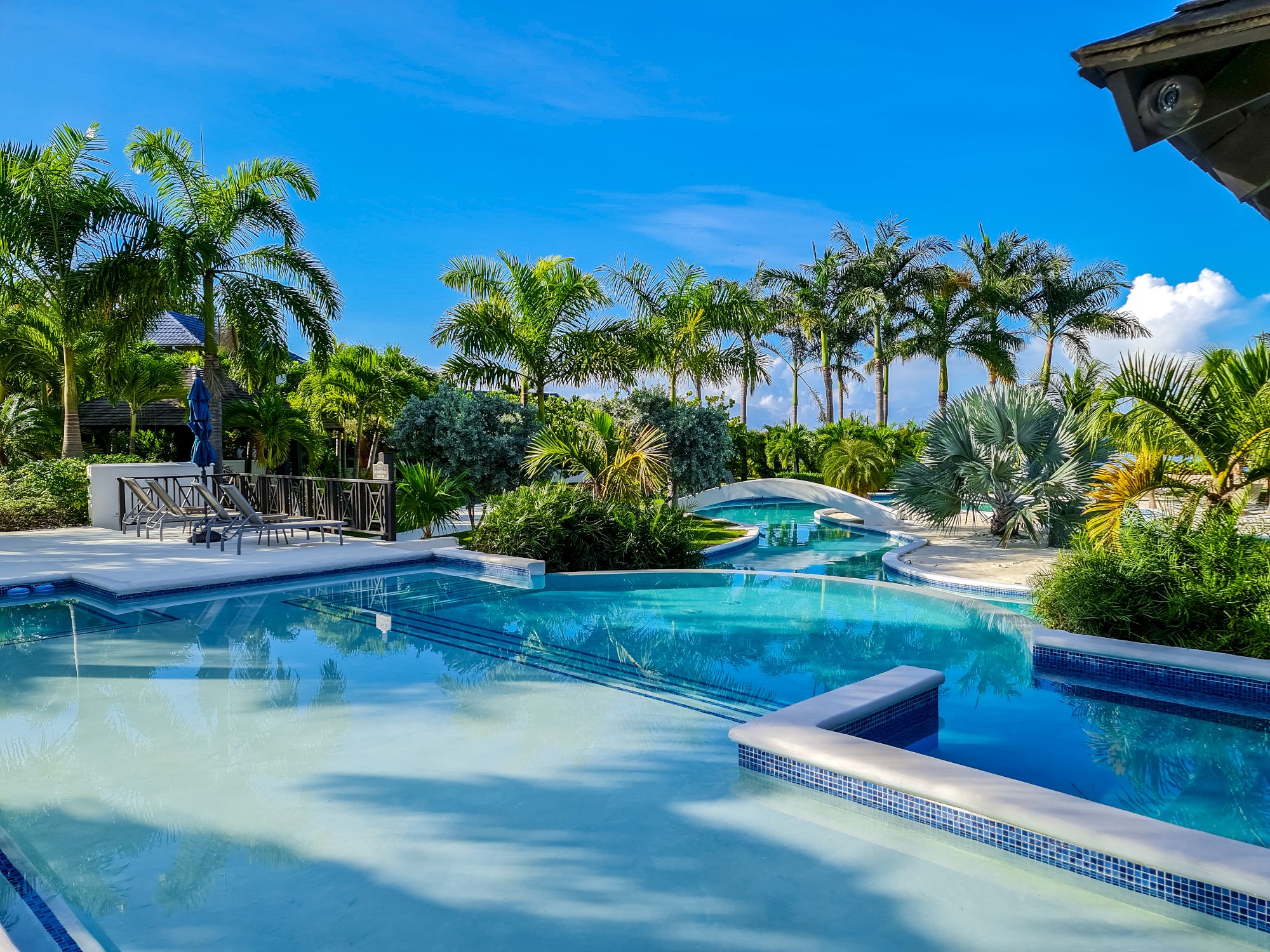 A luxurious resort pool scene with turquoise water, palm trees, lounge areas, and a bright blue sky, suggesting a tropical getaway.