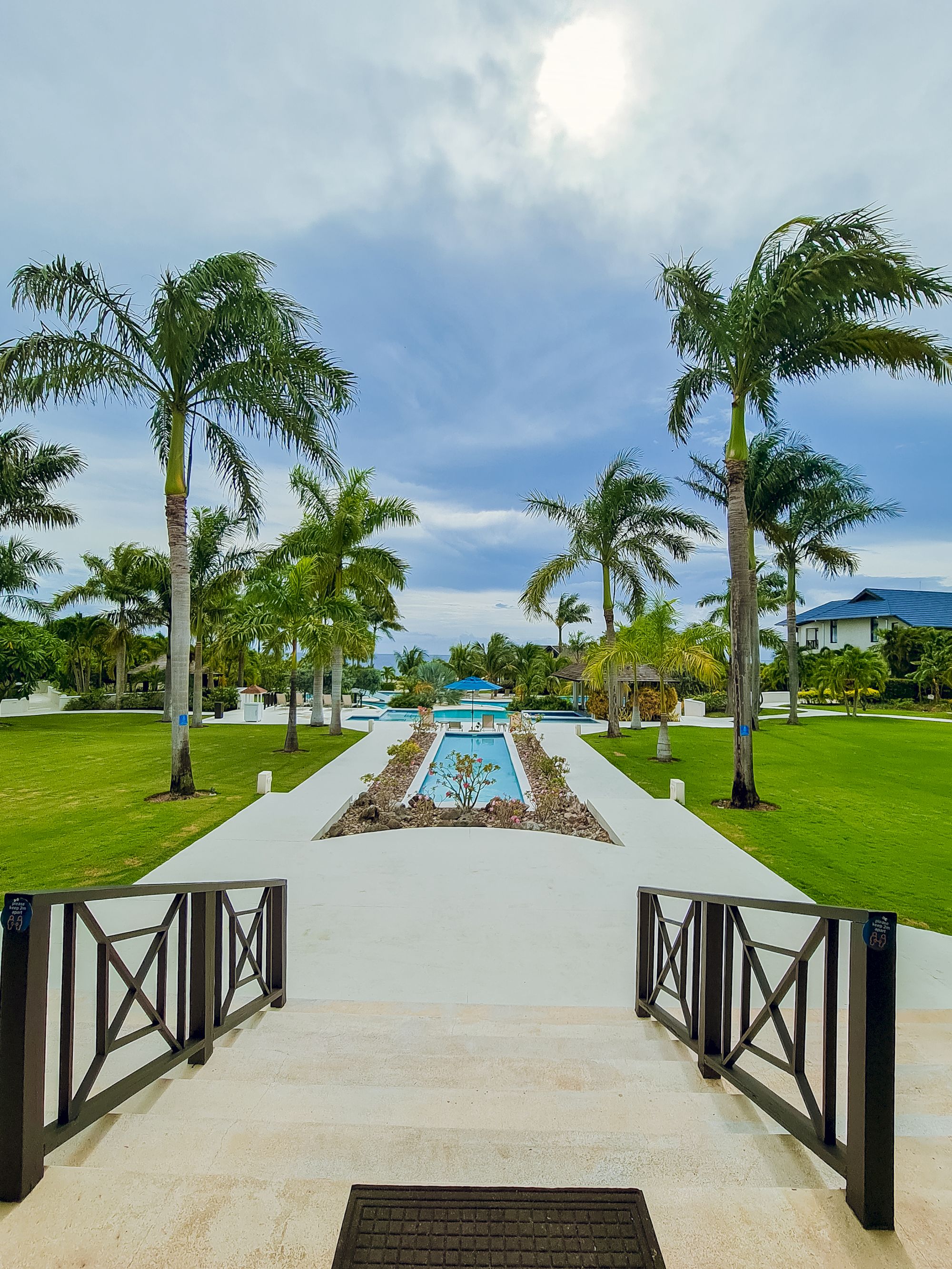 Palm trees line a manicured lawn with a central rectangular pool, a walkway, and a seaside view beyond the ocean horizon, sunny and relaxing.