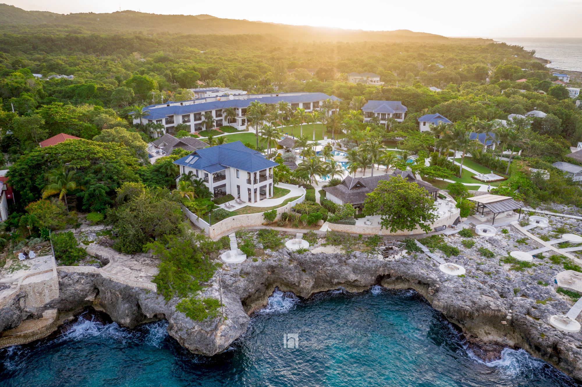 Aerial view of a coastal resort with blue-roofed buildings, lush trees, and rocky shoreline meeting clear blue water at sunset, tranquil and scenic.