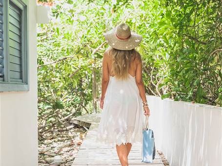 A woman in a white sun dress and hat walks barefoot along a wooden path, carrying a blue bag, shaded by leafy trees.