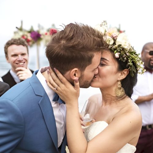 A newly married couple kiss at their outdoor beach wedding, friends and family cheering in the background.