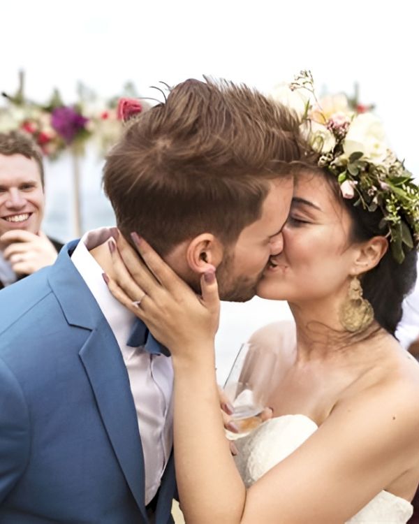 A newly married couple kiss at their outdoor beach wedding, friends and family cheering in the background.