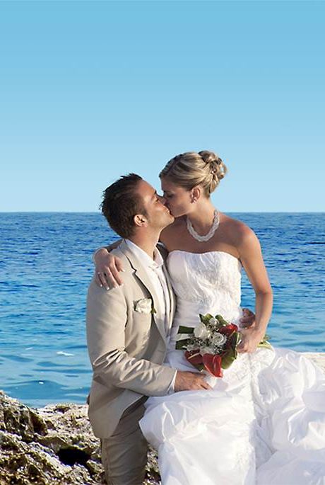 A newlywed couple in wedding attire share a kiss by the rocky shoreline with the sea in the background, posing on a stone staircase.