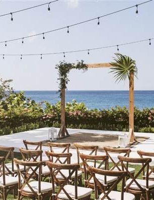Outdoor wedding setup by the sea with string lights, wooden chairs, and a simple wooden arch over a sandy shore backdrop.
