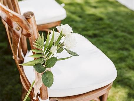 A wooden chair with a white cushioned seat outdoors, decorated with a small green leafy sprig tied to the chair back using twine.