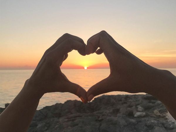 Two hands form a heart shape around a sunset over the ocean, with a rocky shore in the foreground, capturing a romantic seaside moment.