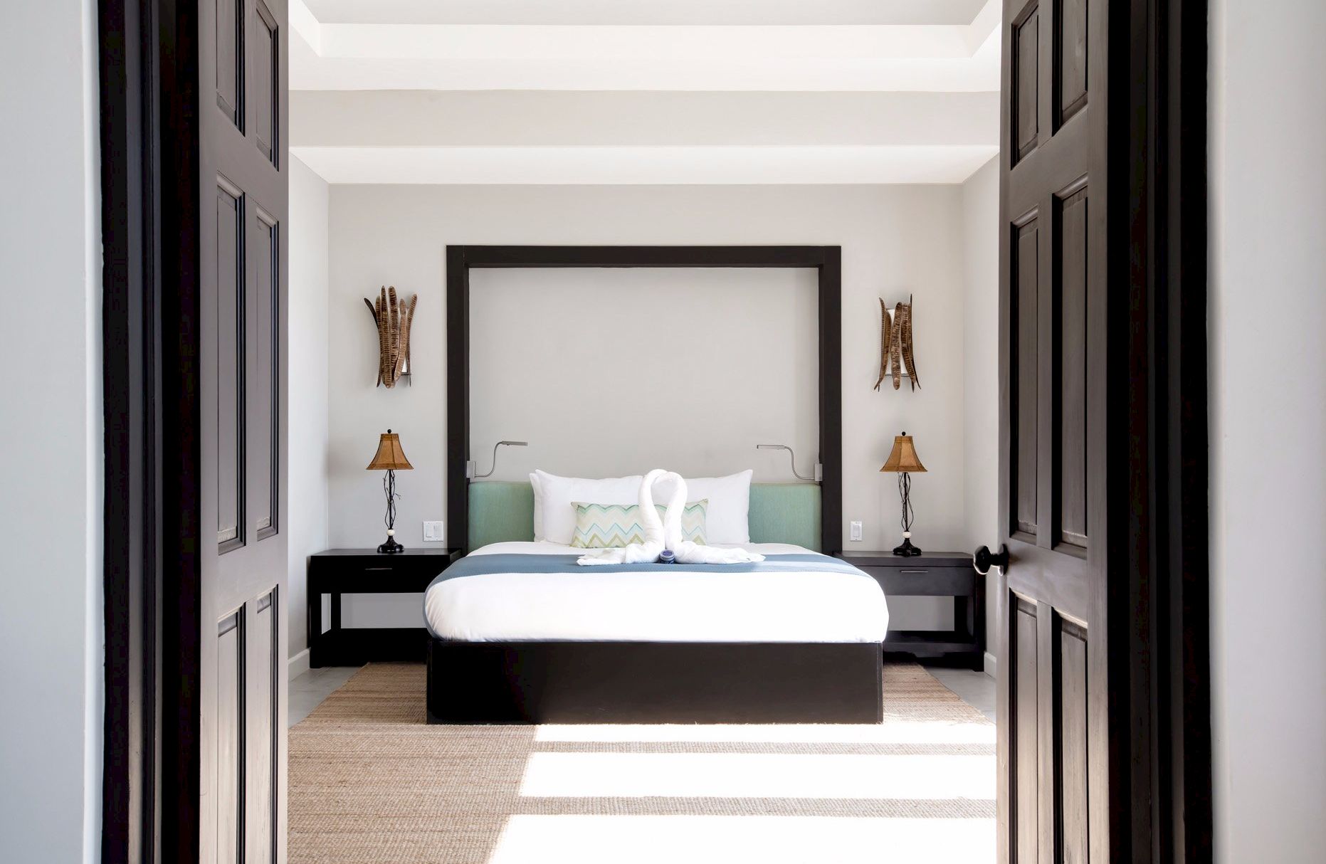 A bright, modern bedroom framed by dark double doors, with a large bed, two black nightstands, lamps, and neutral decor.