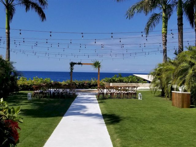 A sunny outdoor wedding setup: a white aisle leads to a wooden arch, string lights overhead, with chairs on a lush lawn and ocean in the distance.