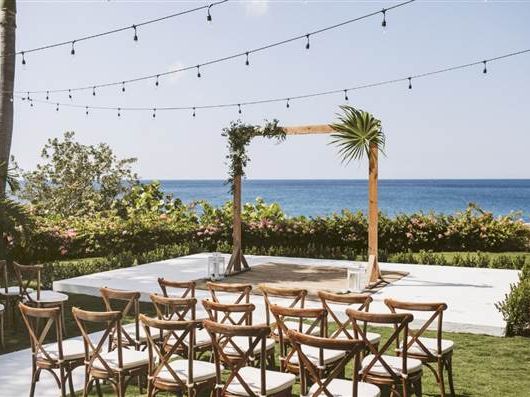 Outdoor beach ceremony setup with wooden chairs, string lights, floral arch, and ocean view in the background, ready for guests.
