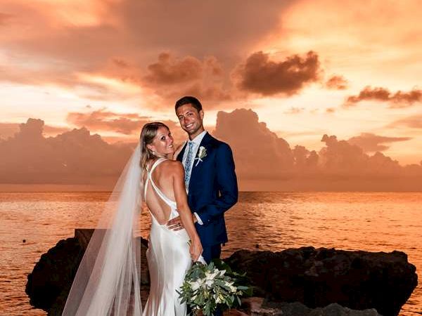 A bride and groom stand on rocks by the sea at sunset, the bride in a long white gown with veil, holding greenery bouquet, smiling at camera.