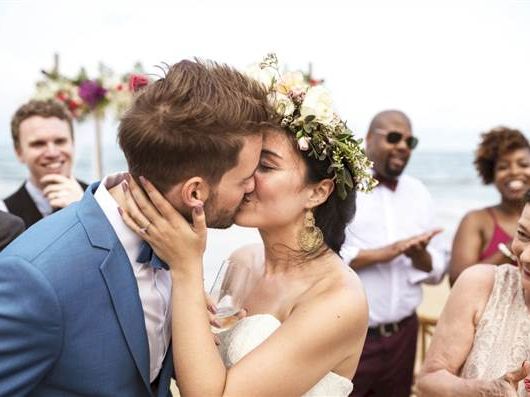 A bride and groom kiss at their outdoor wedding ceremony, guests celebrate in the background by the sea.