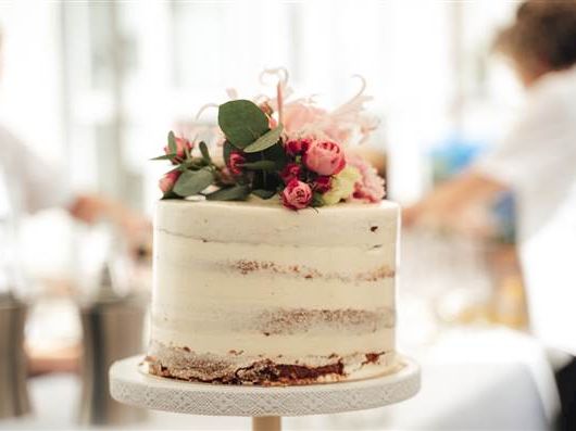 A simple naked cake with light frosting and a floral arrangement topper on a cake stand, set in a bright kitchen.