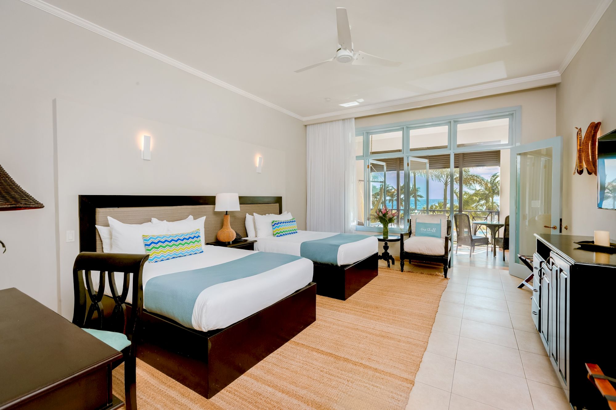 Two large beds with turquoise accents in a bright beachfront hotel room, balcony doors open to a seating area and ocean view, sunny and airy.