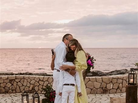A couple hugs on a seaside patio at sunset, sharing a tender moment with lanterns and a stone barrier in the background.