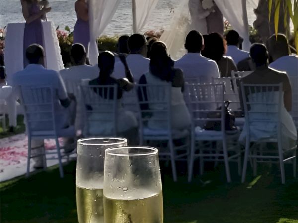 A wedding ceremony outdoors on the beach with a white aisle, seated guests, and two champagne glasses in the foreground.