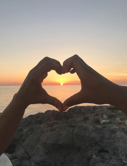 Two hands form a heart frame around a setting sun over the ocean, with a rocky shoreline in the foreground.