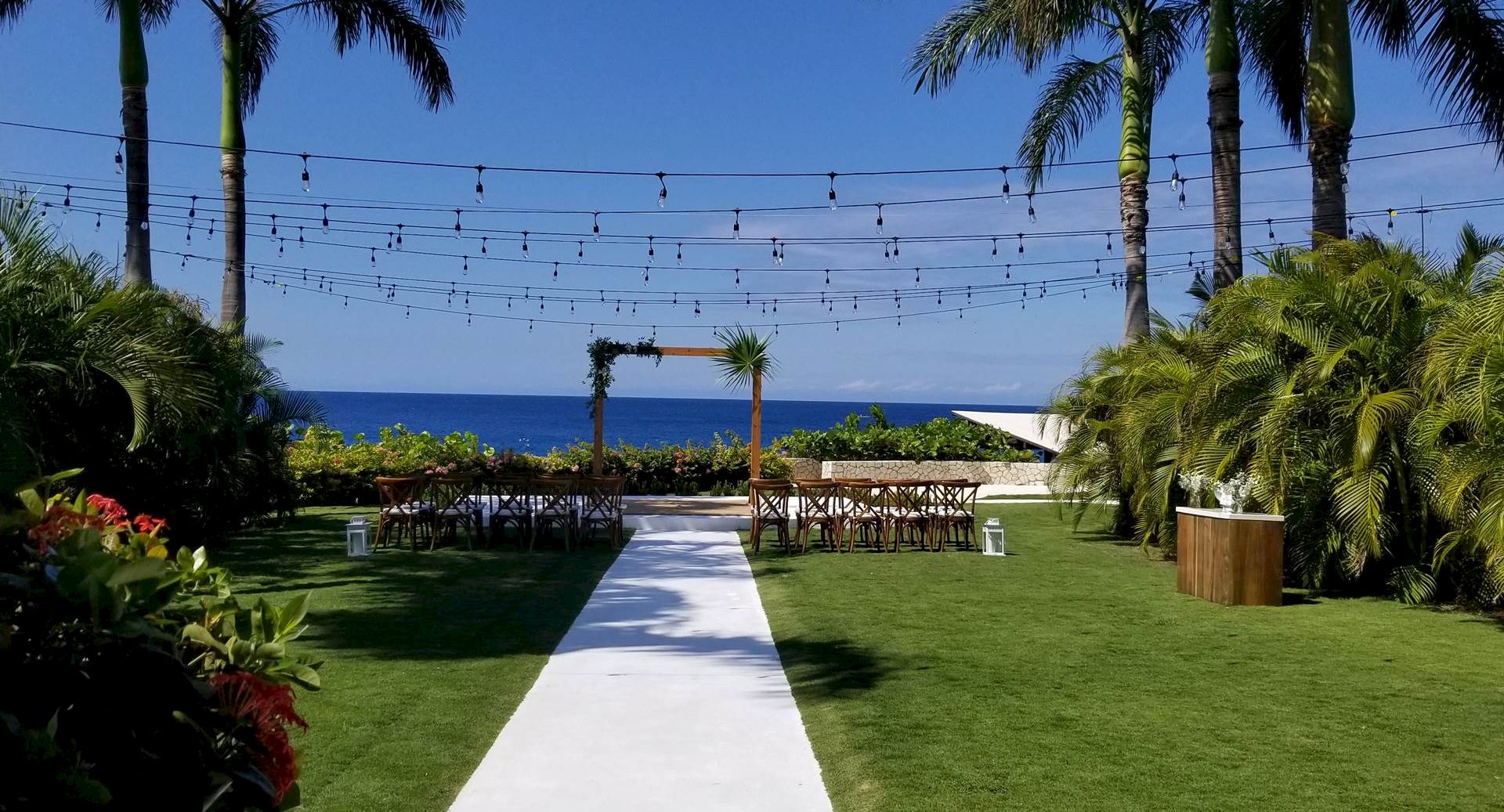 An outdoor wedding setup on a sunny lawn: white aisle runner leading to a wooden arch, string lights overhead, with palm trees and ocean in the background.