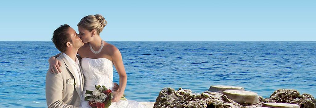 A newlywed couple shares a kiss by the sea, the bride in a white gown and bouquet, with rocky steps and blue ocean in the background.