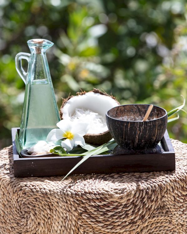 A glass bottle with a narrow neck on a wooden tray, plus a small wooden bowl and a coconut husk, placed on a woven stool outdoors, with greenery in the background.