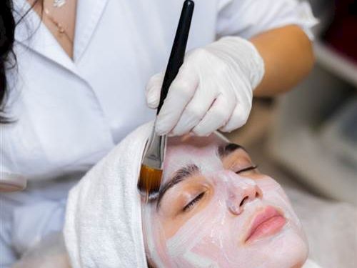A esthetician applies a facial treatment with a brush on a smiling client wearing a headband, a white mask layer ready to set, in a spa setting.