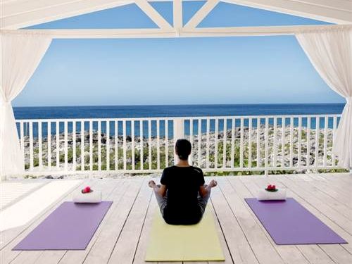 A person sits cross-legged on a yellow yoga mat on a white wooden deck, facing the ocean, flanked by purple mats, under a white-framed cover.