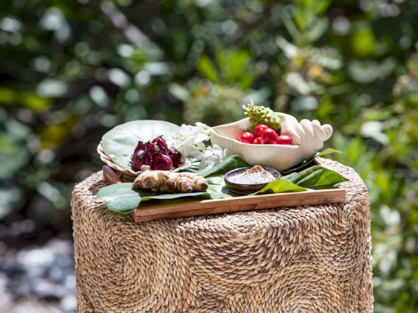 A rustic outdoor food spread with bowls of colorful dishes on a wooden tray&mdash;plus greens and a leaf-wrapped centerpiece on a woven stool, set in a garden.