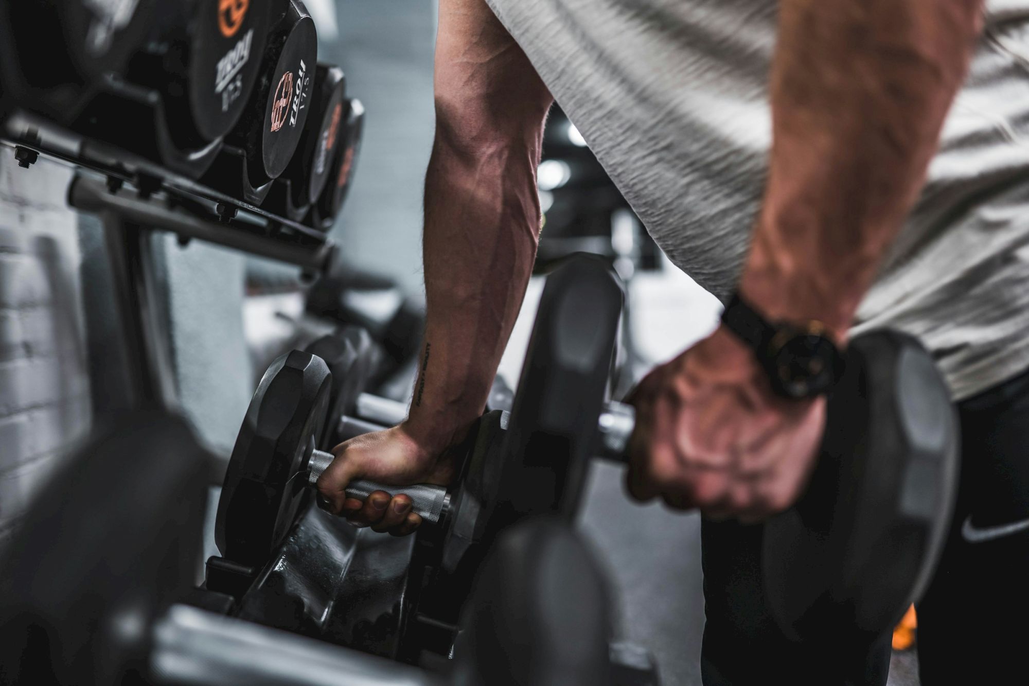 A person gripping a dumbbell near a rack of weights, mid-workout, showing focused effort and a gym setting.