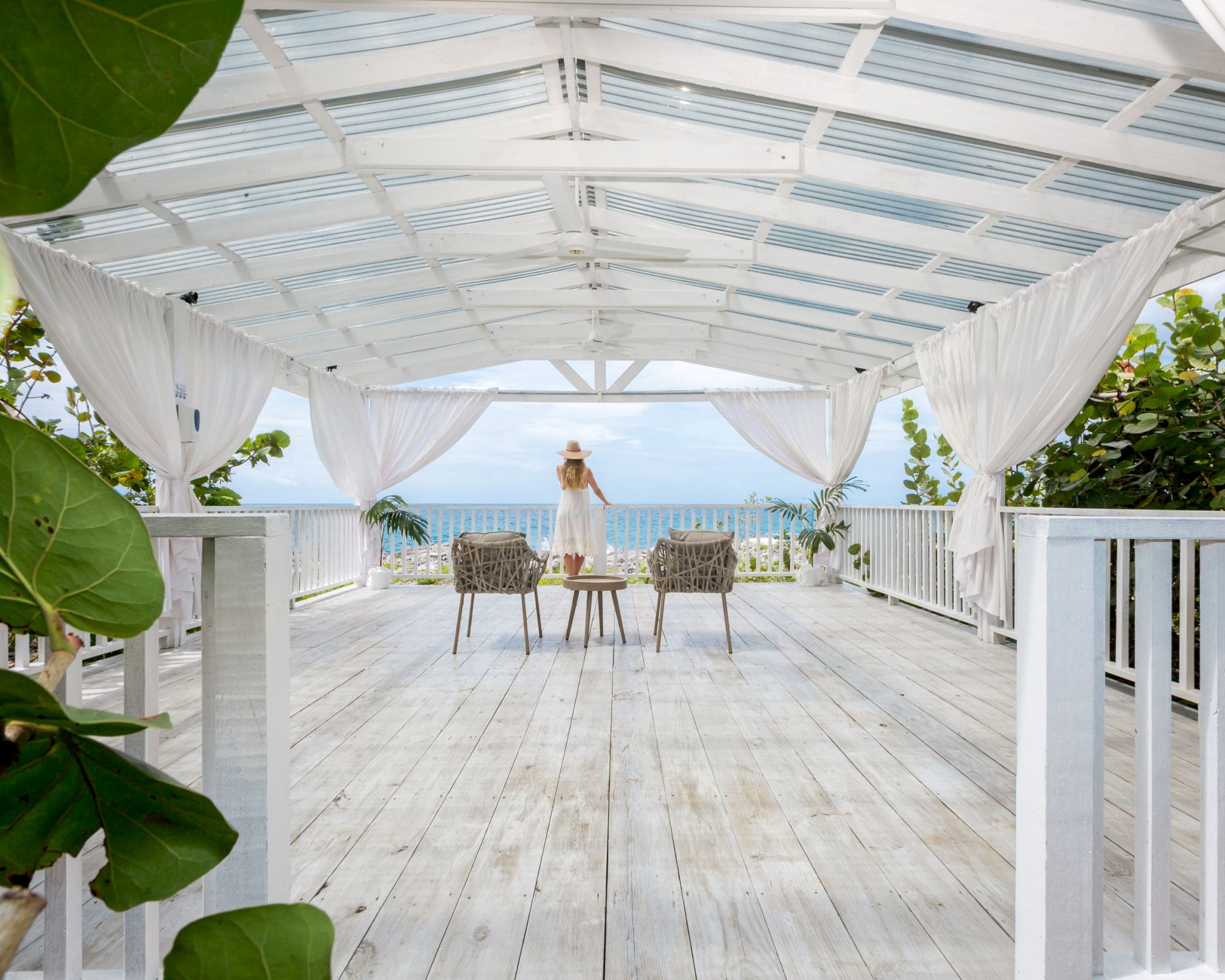 A sunlit beach deck with whitewashed wood, sheer curtains, and lush greenery framing a distant ocean; a person stands near simple chairs.