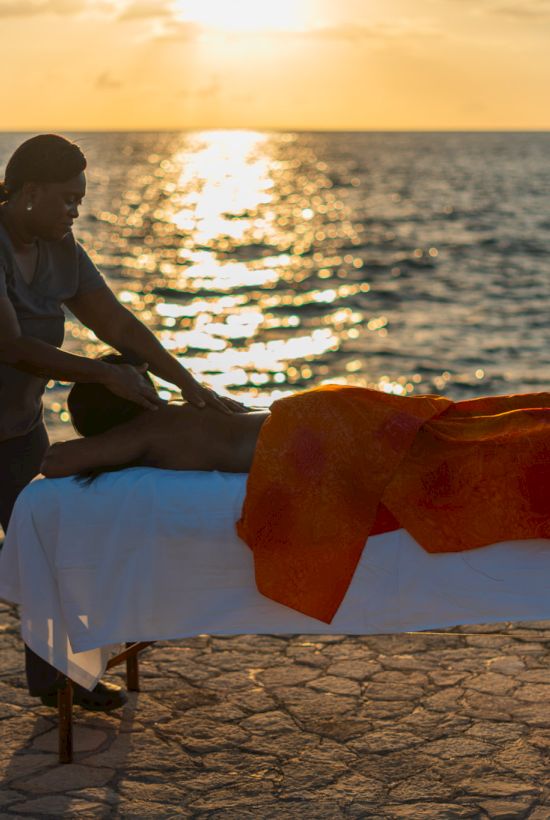 A person giving a massage on a beach at sunset, using a massage table with a white sheet and an orange towel, by the ocean. Top it at 140 characters, always ending the sentence.