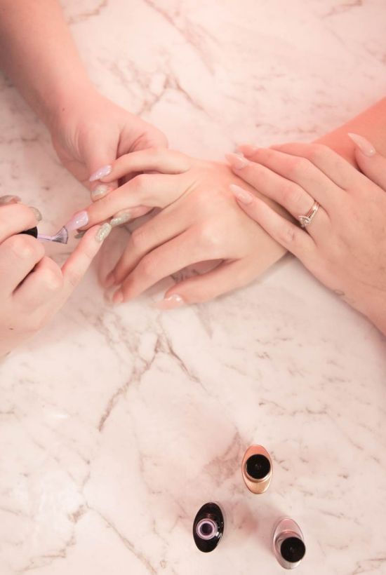 Two people getting a manicure, one hand receiving nail care while the other guides, with nail polish bottles on a marble surface.