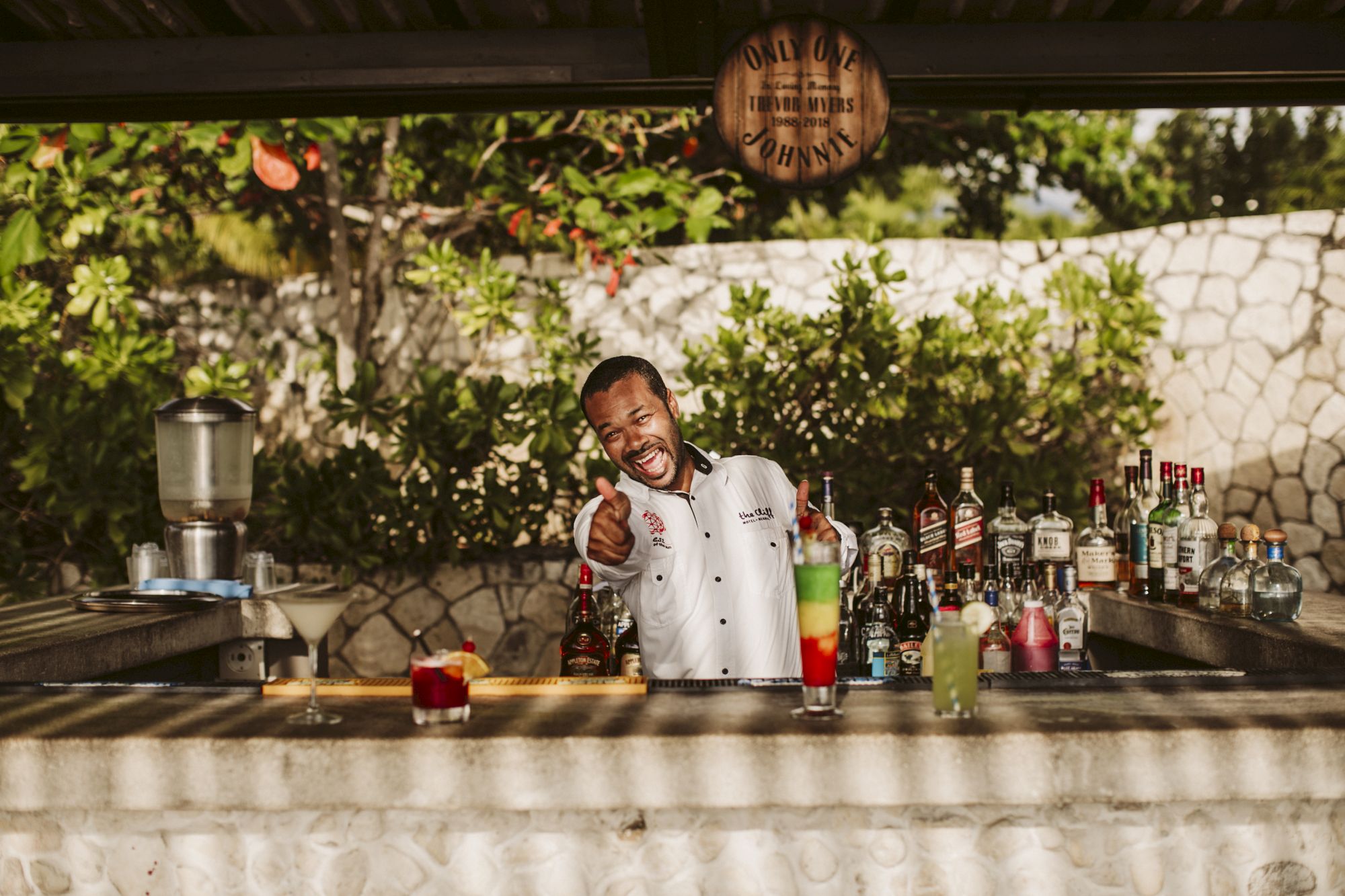 A smiling bartender stands behind an outdoor bar, pointing at the camera with colorful bottles and drinks lined up on the counter.