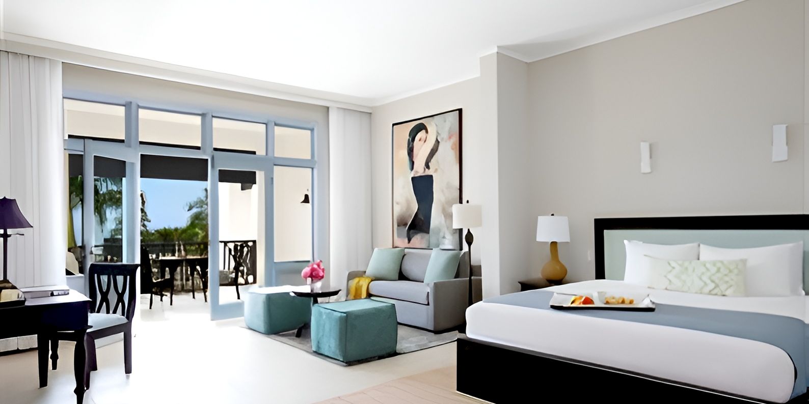 A bright hotel room with a king bed, seating area, and a balcony/patio. Modern decor, soft grays and whites, with a large window and framed art.