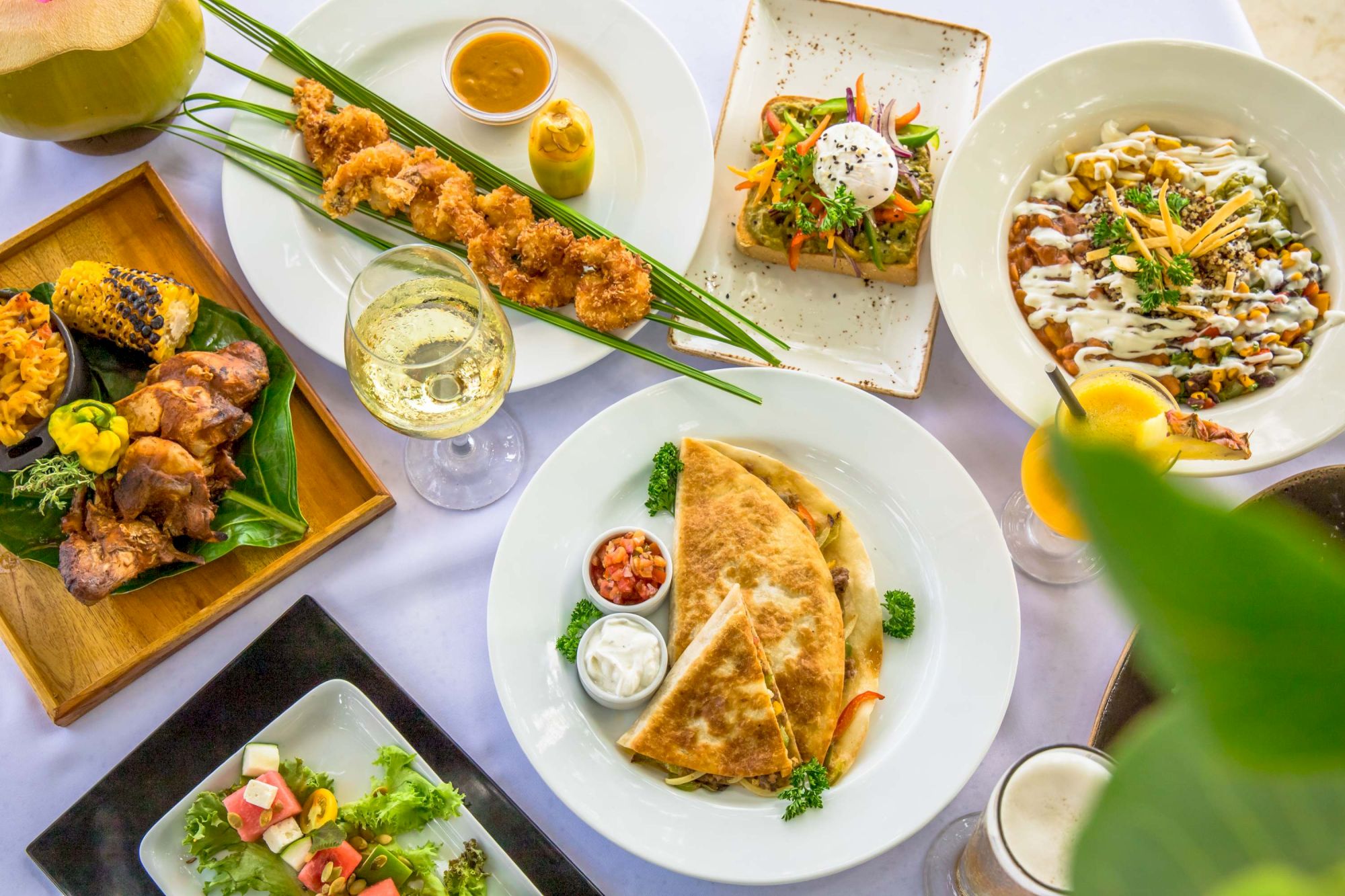 A vibrant spread of dishes: nachos, flatbread topped with greens, a bowl of soup, fried skewers, salads, a quesadilla, and drinks, all on a bright table.