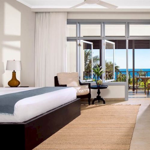 A bright hotel room with a large bed, nightstand, chair, and a view of a balcony overlooking the ocean and palm trees.