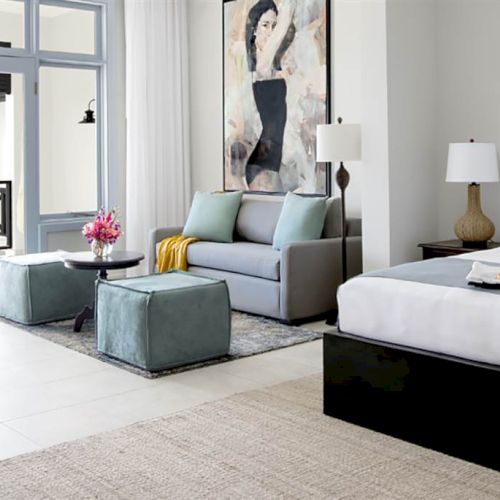 A bright hotel-style hotel room with a large bed, seating area, glass doors to a balcony, modern decor, and a tray of fruit on the bed.