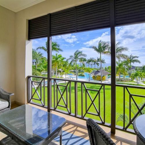 A balcony dining area with wicker furniture overlooks a sunny lawn and a pool-like view framed by palm trees.