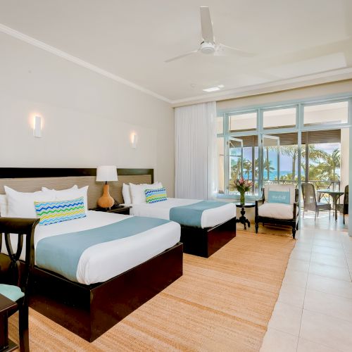 A bright hotel room with two queen beds, a seating area, a balcony view, and a mini-fridge/desk setup in a modern, tropical-style suite.