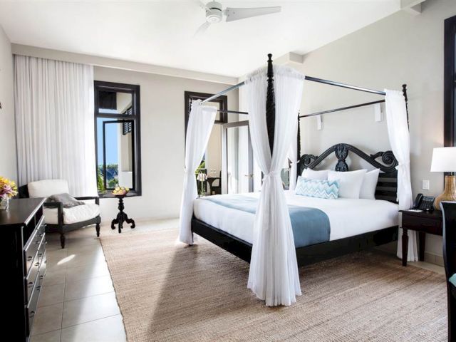 A luxurious hotel room with a four-poster bed, sheer curtains, a seating area, and large windows letting in natural light.