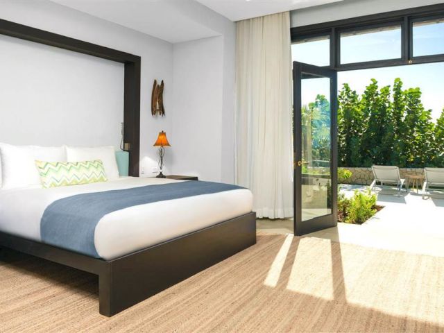 A modern hotel room with a dark wood bed, white bedding, mint-green accents, and a sliding glass door opening to a sunny patio and pool area, ending here.