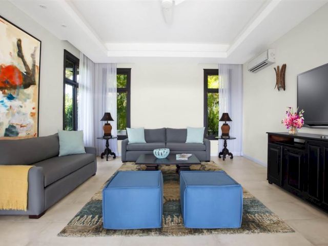 A bright, modern living room with two gray sofas, blue ottomans, a central wooden coffee table, a rug, artwork, and a black TV console with a flat-screen TV.