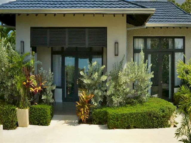 A modern single-story house with large glass doors, tropical landscaping, tidy hedges, and potted plants lining a sunny entrance.