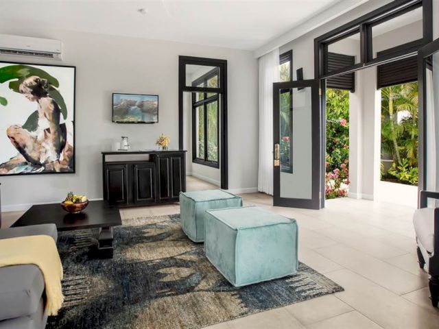A modern living room with large glass doors, a dark coffee table, mint green ottomans, a black cabinet, and tropical wall art.