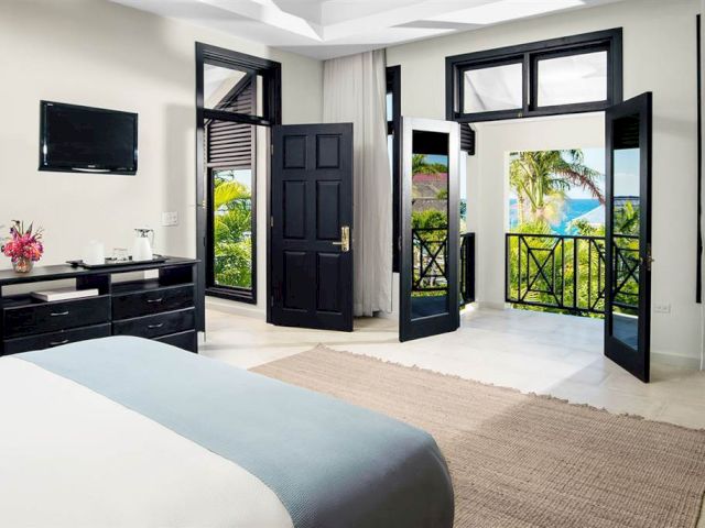 A bright hotel room with a bed, dresser, and large glass doors opening to a balcony with a garden view.