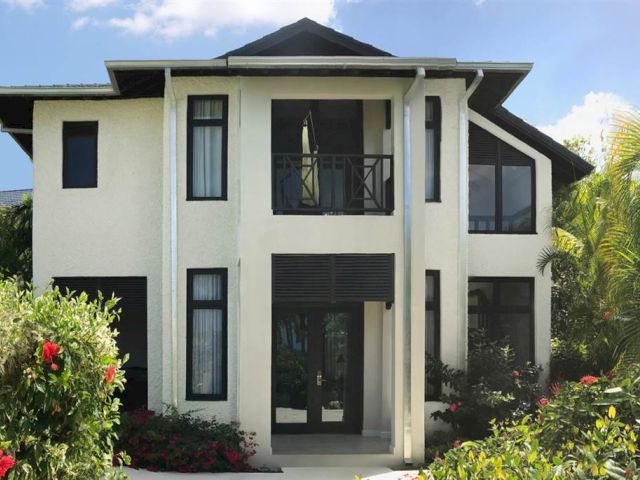 A modern two-story house with large windows, a central front door, and tropical landscaping surrounding it.