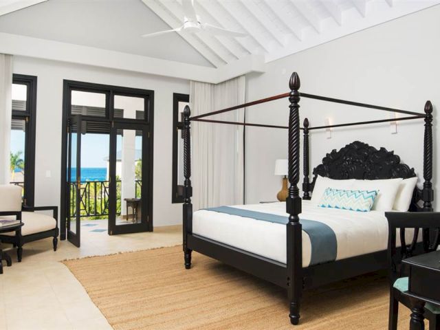 A luxurious dark wood four-poster bed with white and blue bedding sits in a bright, airy hotel room with a balcony view.