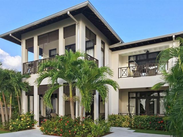 A modern two-story house with white walls, dark trim, and large windows, surrounded by palm trees and tropical landscaping, sunny day.