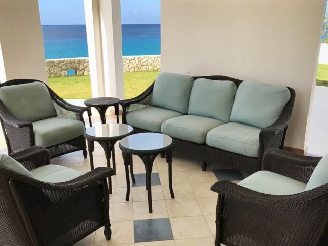 A covered outdoor seating area with wicker chairs and a sofa set around small round tables, overlooking a lawn and blue sea beyond.