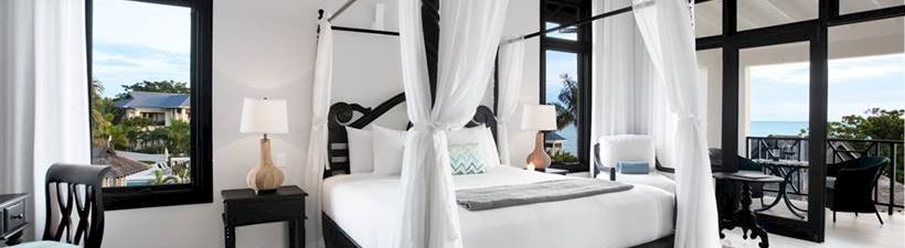 A luxurious white-canopy bed anchors a bright, airy bedroom with black-framed windows, a woven rug, and a balcony view&mdash;elegant coastal chic.