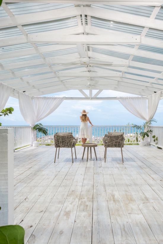 A person stands at a bright, airy white wooden deck pavilion overlooking the ocean, with chairs and a table set under draped curtains.